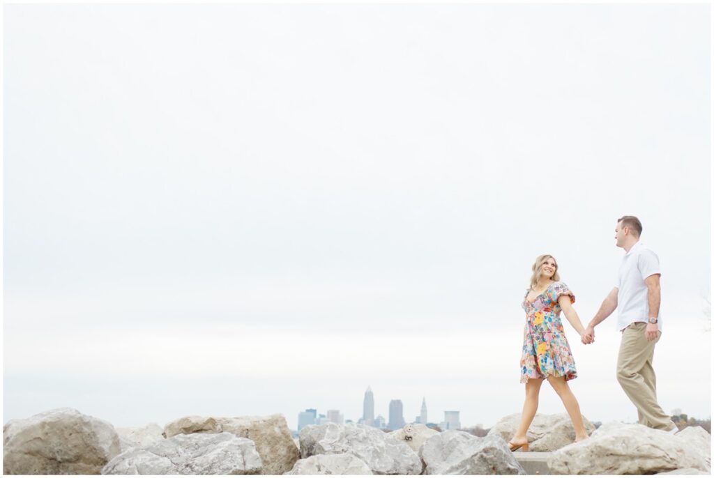 An engaged couple posing at Edgewater park for their engagement session with the downtown cleveland skyline behind them