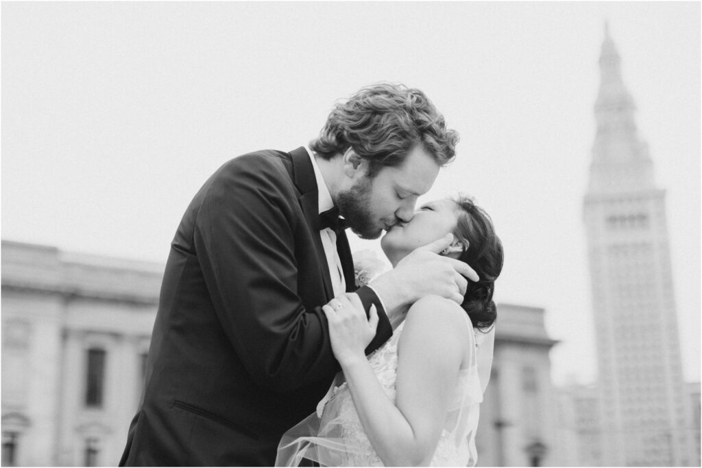 A black and white photo of a bride and groom kissing with Terminal Tower in the distance in Cleveland Ohio.