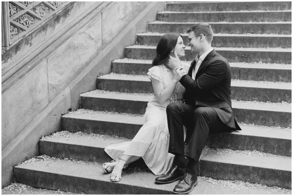A black and white photo of a couple sitting on steps in Central Park for their New York City engagement session